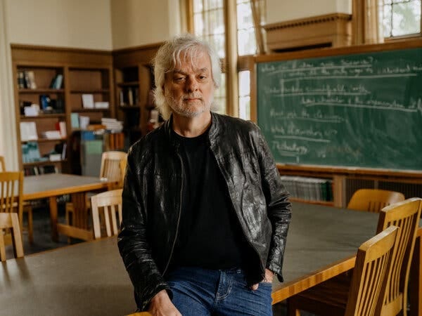 A portrait of David Chalmers, wearing jeans and a leather jacket, leaning against a table in a room with a blackboard, desks and bookcases.