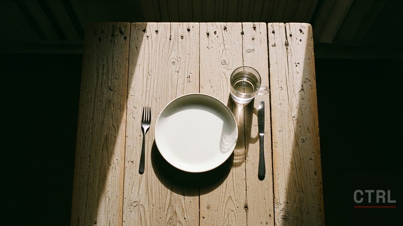 Overhead view of a simple place setting on a worn wooden table with a single plate and coffee mug in natural window light, suggesting quiet appreciation for life's essentials."
