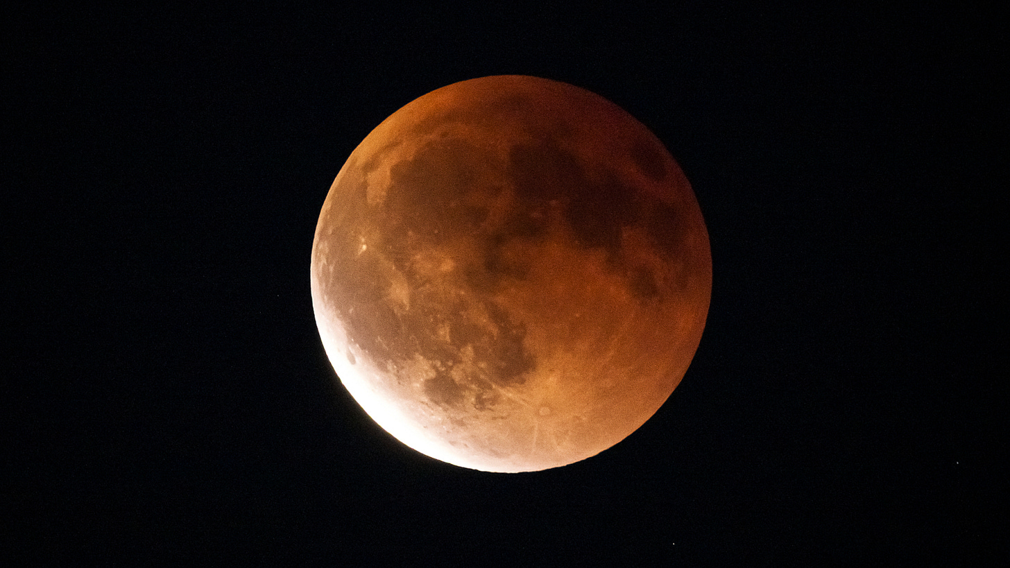 A yellow-orange eclipsed moon is pictured against a dark sky, with dark features known as lunar maria marking its surface along with the shapes of bright impact craters. The lower left part of its disk is brighter than the rest of its surface. A yellow-orange eclipsed moon is pictured against a dark sky, with dark features known as lunar maria marking its surface along with the shapes of bright impact craters. The lower left part of its disk is brighter than the rest of its surface.