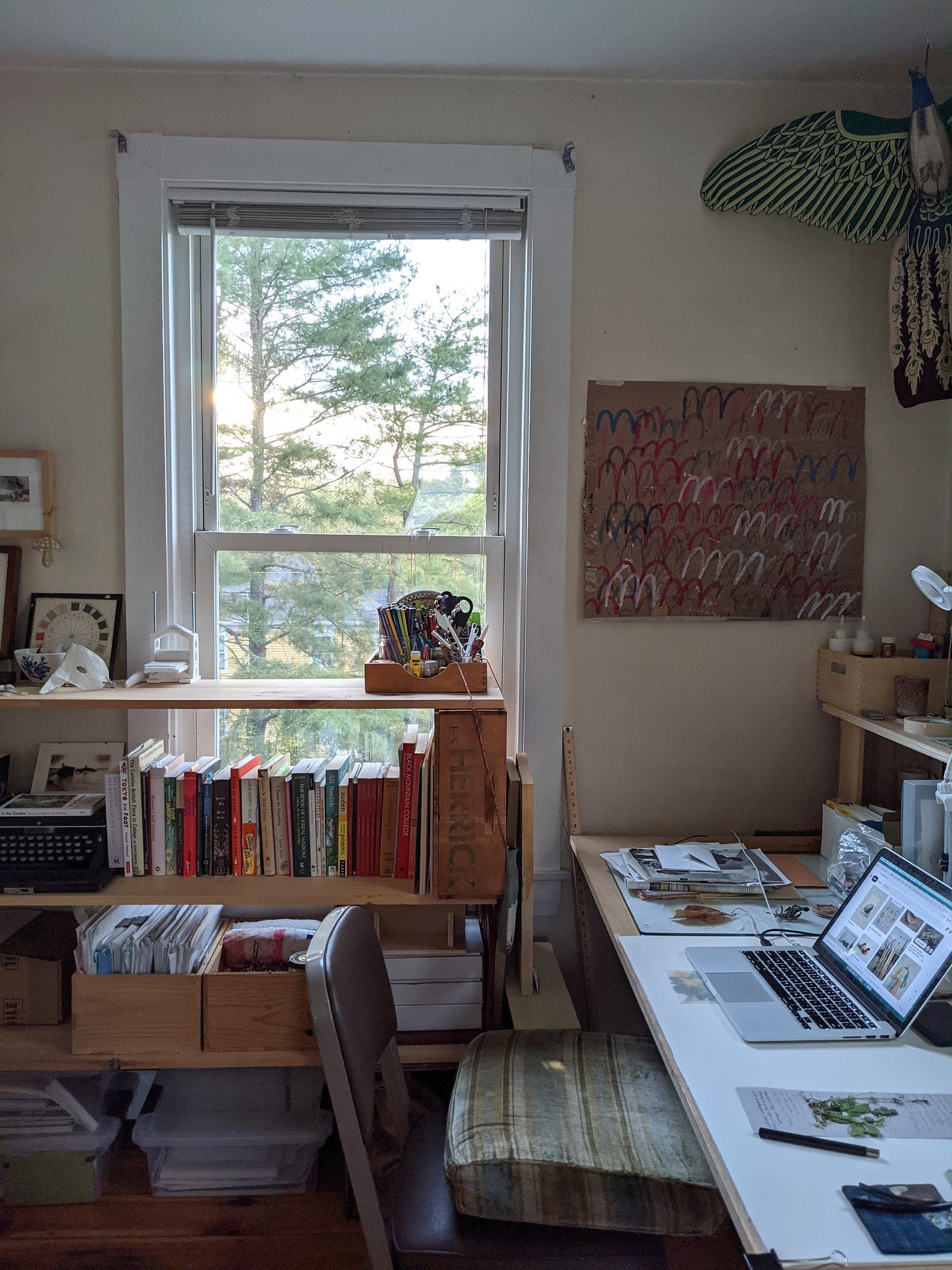 a room with bookshelves made of boards and boxes, a desk made of a board and trestles, a chair. books, pictures on the wall, a kite shaped like a peacock.