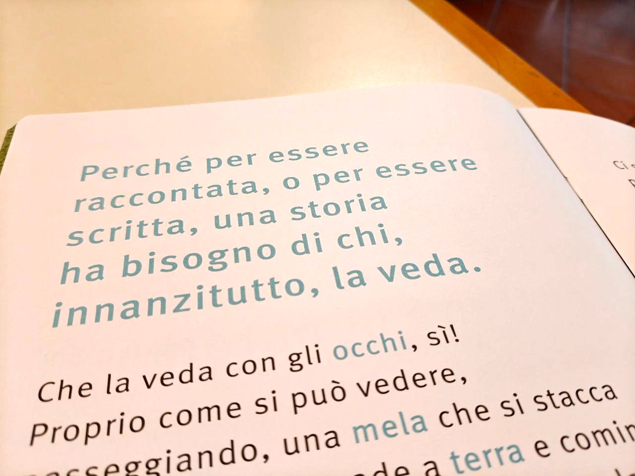 Una pagina della parola LIBRO delle Scatoline di effequ, con questa scritta: "Perché per essere raccontata, o per essere scritta, una storia ha bisogno di chi, innanzitutto, la veda". La pagina è appoggiata su un tavolo di legno bianco sporco e nell'angolino in alto a destra si intravede un pezzetto di pavimento in cotto.