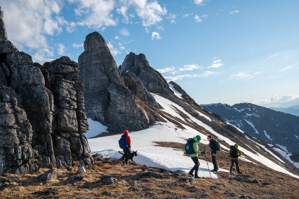 Four hikers and a dog walk along a tundra-and-snow ridge past tall granite spires in warm evening light.