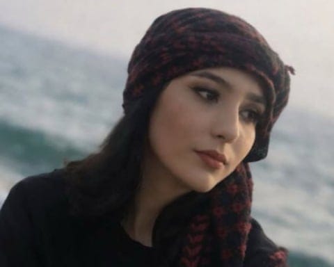 A young Middle Eastern woman, in a patterned headscarf, looks off camera with the sea blurred behind her