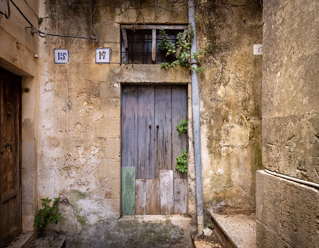 A weathered blue-gray wooden door patched with odd planks in a crumbling limestone wall in Modica Bassa, with blue-and-white house numbers and a barred window above. A weathered blue-gray wooden door patched with odd planks in a crumbling limestone wall in Modica Bassa, with blue-and-white house numbers and a barred window above.