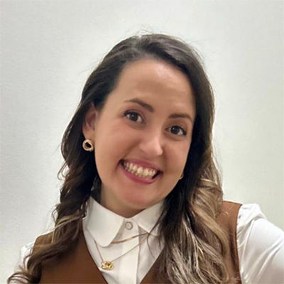 Retrato de uma mulher sorrindo para a câmera, em um ambiente interno com parede clara ao fundo. Ela tem pele clara, cabelos castanhos longos e ondulados, usa brincos e colares dourados, camisa branca de mangas compridas e colete marrom com botões. Está levemente inclinada para a direita, com uma das mãos apoiada na cintura, transmitindo uma expressão alegre e acolhedora.