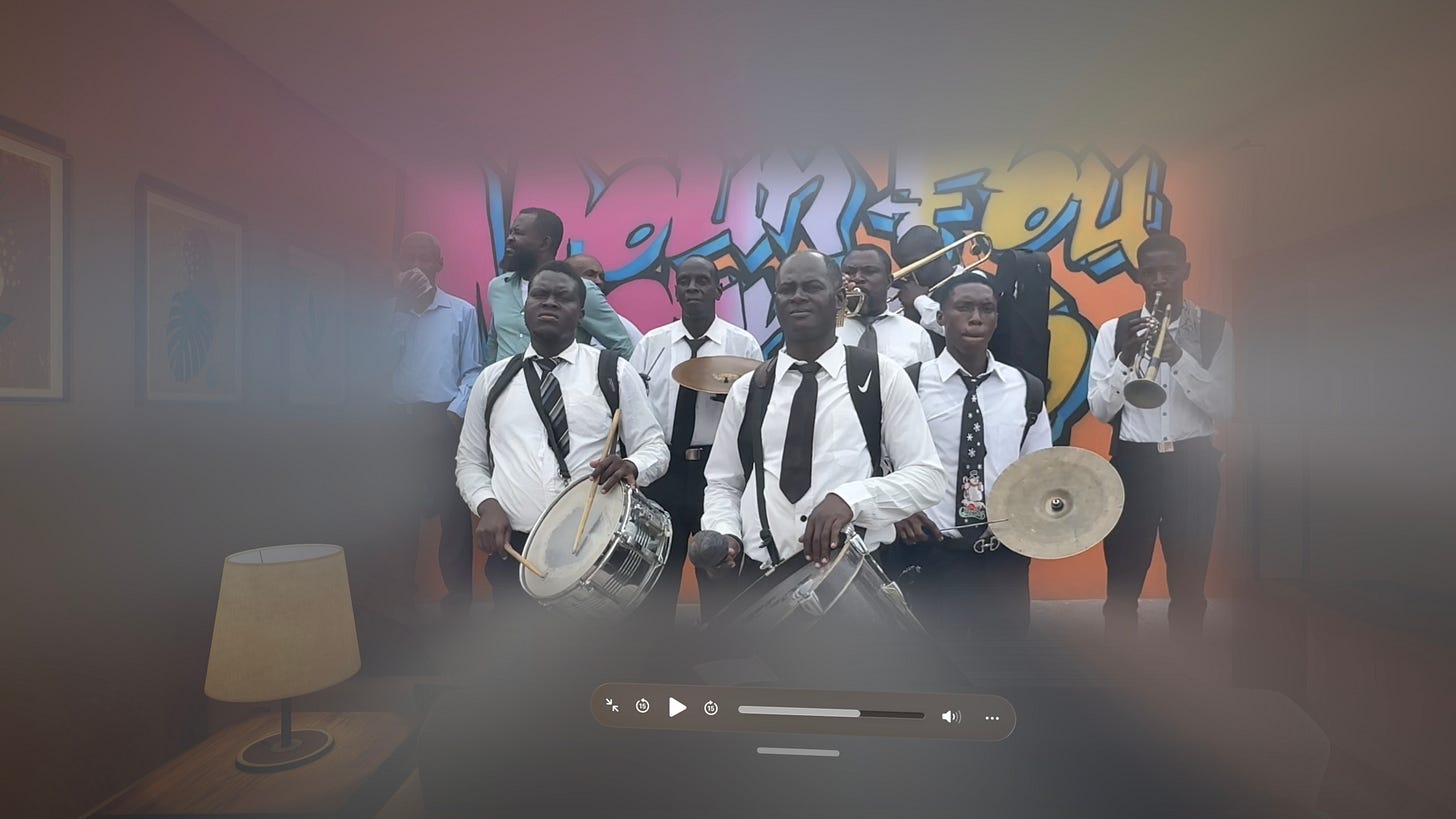 A brass and percussion band of approximately eight musicians performs in front of a vibrant graffiti mural in orange, pink, and blue. The musicians in the foreground wear white dress shirts, dark ties, and suspenders, and are playing snare drums and cymbals, while brass players with a trombone and trumpet stand behind them. The image is a paused video frame, shown with a media player control bar visible along the bottom.