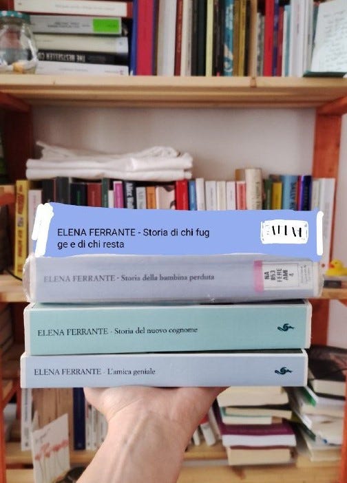 Una mano regge tre libri. Un quarto è disegnato, in cima agli altr tre. Sono i quattro libri, in tinte color pastello, della saga dell'Amica Geniale di Elena Ferrante. Sullo sfondo c'è una liberia piena di libri e altri piccolo oggetti.