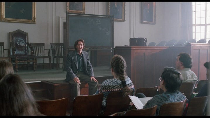 Timothy Hutton as Thad Beaumont sits casually on a desk, speaking to college students in a wood-paneled classroom with a chalkboard behind him. The word “DUALITY” is faintly visible on the board.