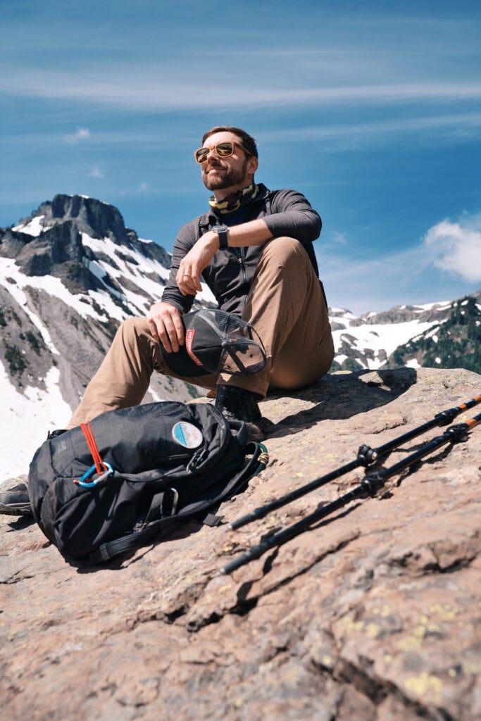 AJ on a mountain with a mountain behind him holding his black hiking had and his camera bag in front of him