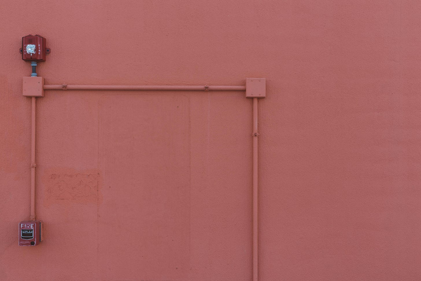 Surface-mounted electrical conduit and a red fire alarm running across a bare salmon-pink exterior wall. Surface-mounted electrical conduit and a red fire alarm running across a bare salmon-pink exterior wall.