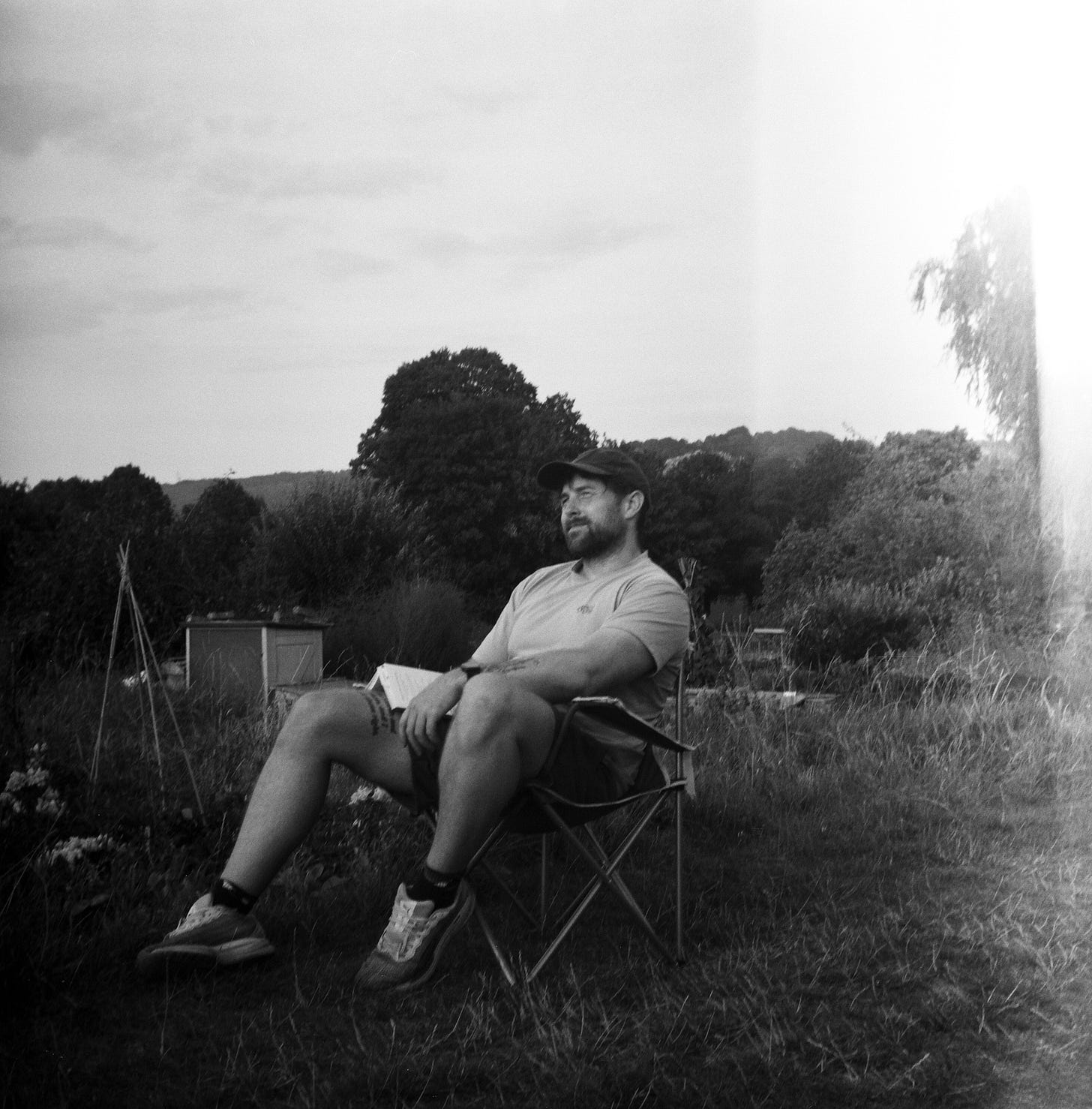 A man sits on a camping chair, looking relaxed. A book is balanced on his leg, covering part of a tattoo, while he looks off into the distance. He's positioned next to a vegetable plot. A man sits on a camping chair, looking relaxed. A book is balanced on his leg, covering part of a tattoo, while he looks off into the distance. He's positioned next to a vegetable plot.