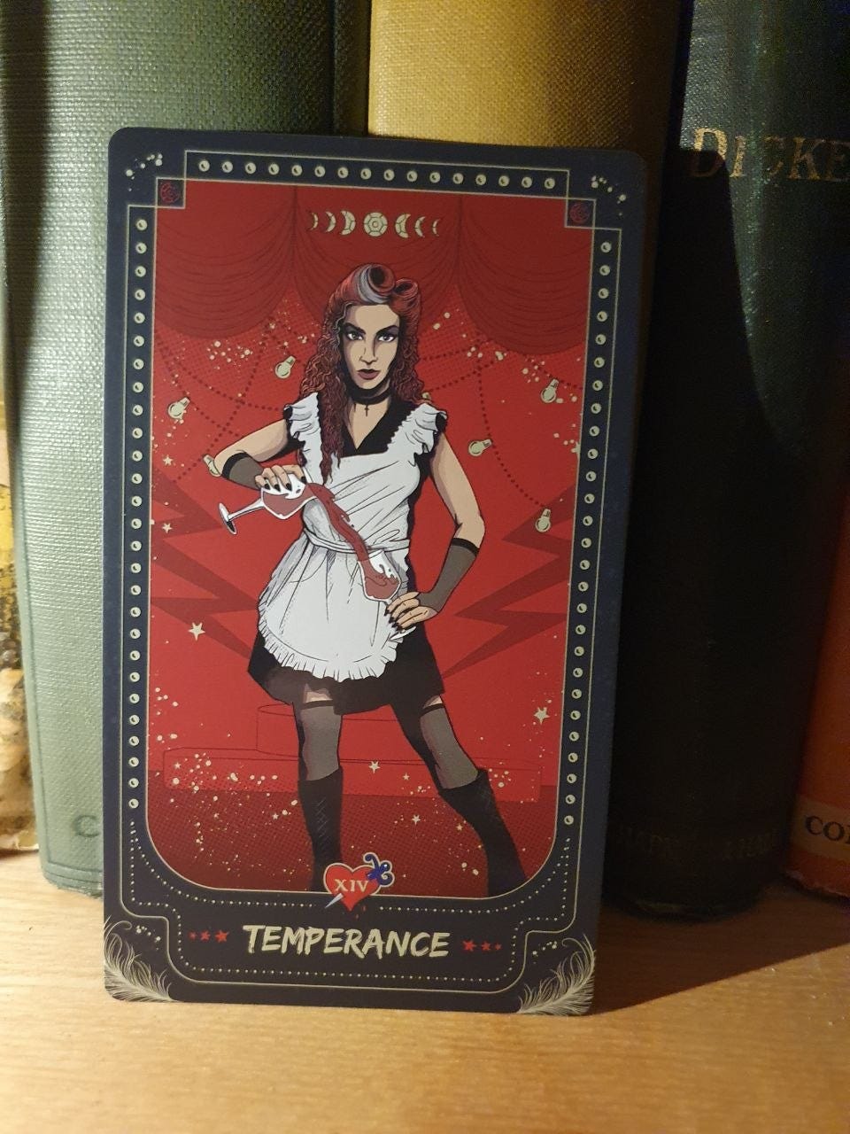 Temperance tarot card, XIV, featuring a theatrical red-haired figure in a black-and-white dress holding a wine glass against a red stage backdrop decorated with stars and moon phases. Temperance tarot card, XIV, featuring a theatrical red-haired figure in a black-and-white dress holding a wine glass against a red stage backdrop decorated with stars and moon phases.