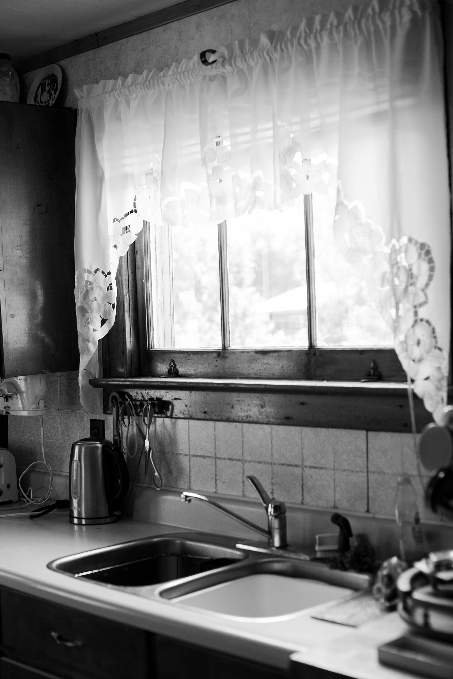 Black and white photo of the kitchen sink. Window above with lace half curtains.