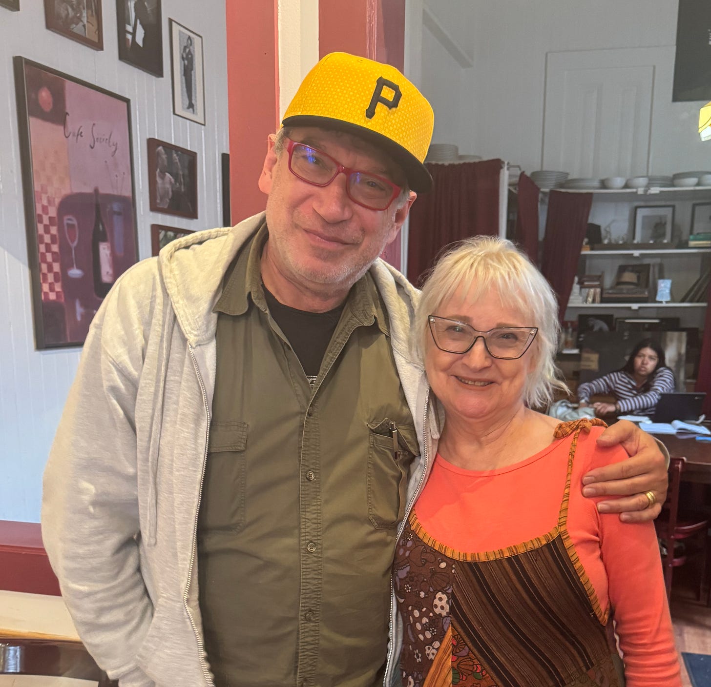 Gary, wearing a yellow Pittsburgh Pirates cap and red glasses, with his arm around Melanie, in Cafe Society, a Half Moon Bay coffeehouse. Gary, wearing a yellow Pittsburgh Pirates cap and red glasses, with his arm around Melanie, in Cafe Society, a Half Moon Bay coffeehouse.
