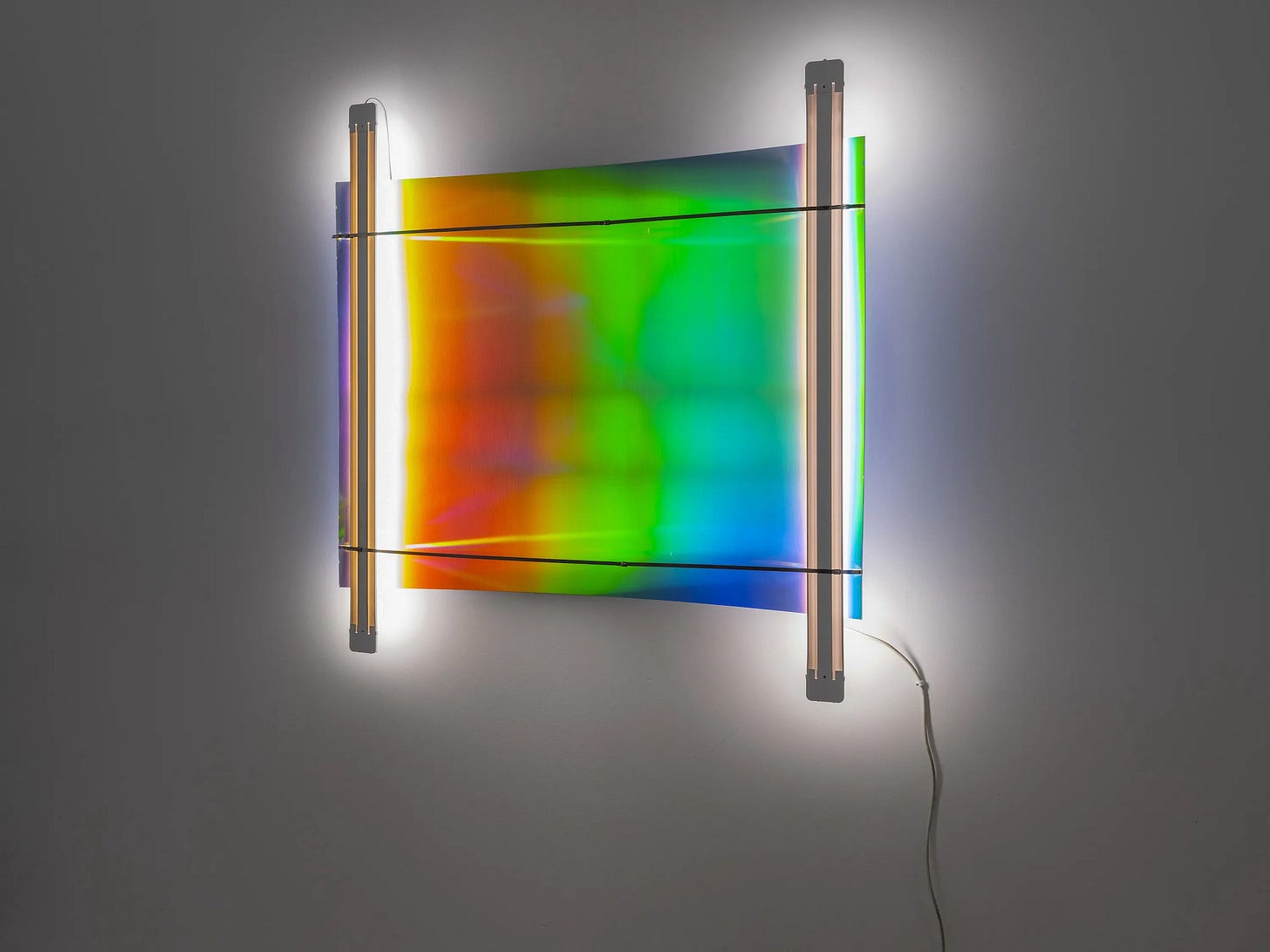 A rectangle of shifting bright colours sits against a darkened gallery wall, lit from either side. A rectangle of shifting bright colours sits against a darkened gallery wall, lit from either side.
