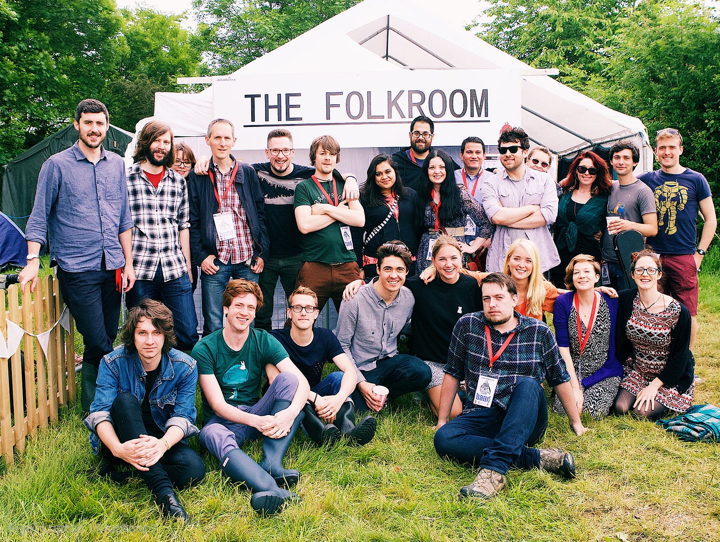 A photo of musicians outside a marquee. Everybody is positioned semi-orderly, as though in a school photo. A banner on the marquee reads 'The Folkroom'. A photo of musicians outside a marquee. Everybody is positioned semi-orderly, as though in a school photo. A banner on the marquee reads 'The Folkroom'.