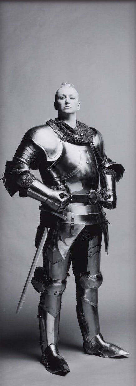1) A woman stands confidently in full medieval armor, featuring polished metal plates and chainmail. The grayscale image has a serious and heroic tone. The person is white with short hair, and she is standing with her armored hands on her hips. There is a sword attached to her right hip. She does not have a helmet, but she does have metal boots. 2) A brown woman in medieval armor with a confident pose, standing against a plain backdrop. The black-and-white image conveys a serious, historical tone. She has shoulder-length dark hair, and she is wearing a white long-sleeve undershirt, a vest, a belt with large buckle, dark pants, and dark boots. On her right arm, there is a sign that includes the names: 'Queen Christina, Natalie Barney, Gluck, Romaine Brooks, and Dorothy Arzner', which are famous historical lesbians and/or artists. She is also holding a dagger in front of her. Her vest is textured and her pants are chainmail.