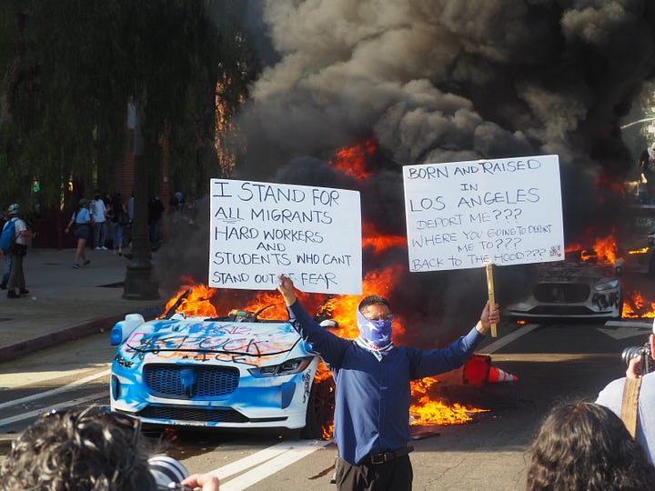 Protesters gathering around three burning and vandalized Waymo rideshare vehicles in Downtown Los Angeles.