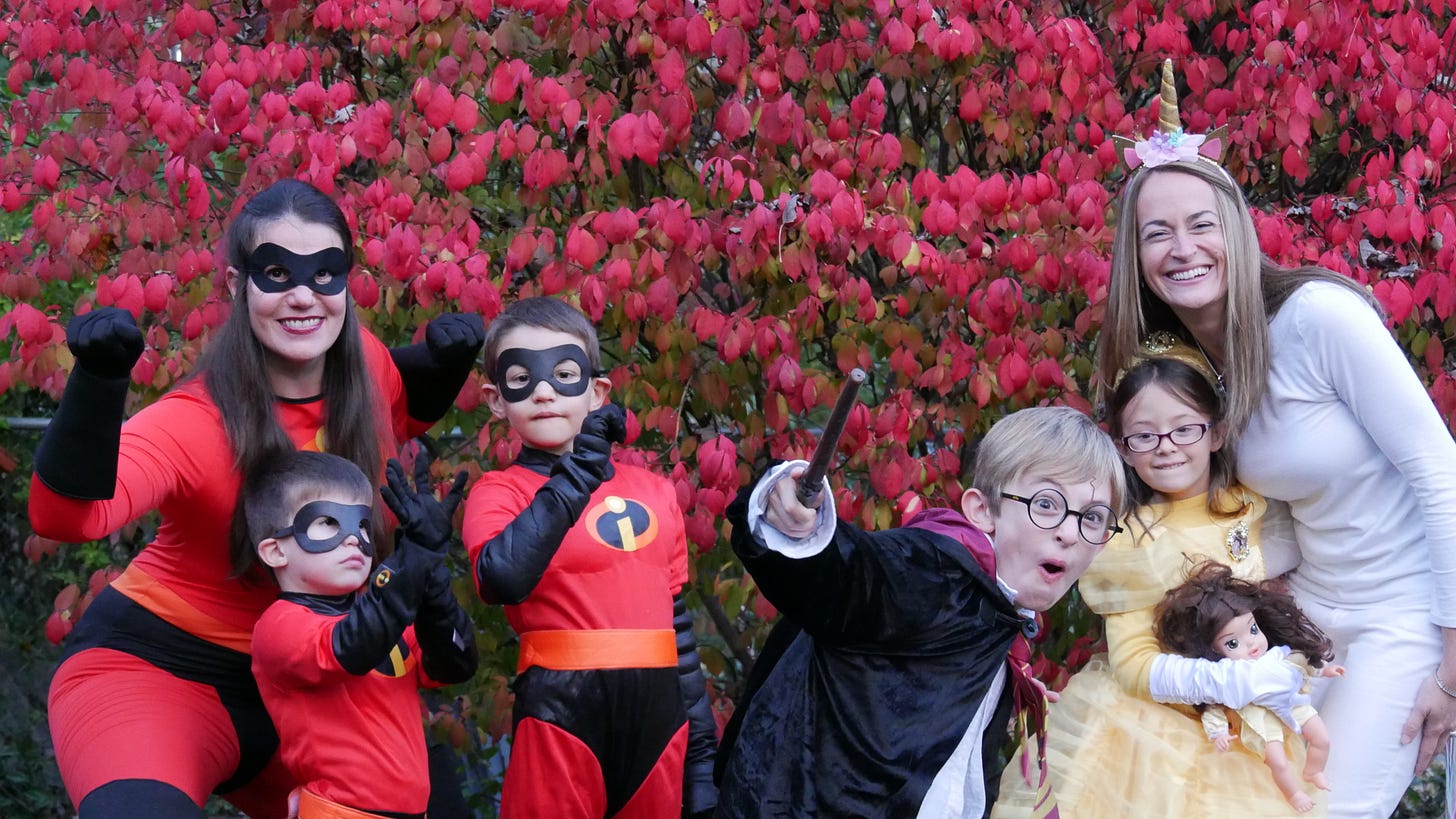 There is a woman and two boys dressed like the family from the Incredibles with red outfits and black masks, a boy dressed like Harry Potter with a robe, glasses, and a wand, a girl dressed like Belle from Beauty and the Beast with a yellow dress, and a woman dressed like a unicorn in white and with a horn and ears. They all stand together smiling at the camera with red leaves behind them. It is a late fall, cloudy afternoon. There is a woman and two boys dressed like the family from the Incredibles with red outfits and black masks, a boy dressed like Harry Potter with a robe, glasses, and a wand, a girl dressed like Belle from Beauty and the Beast with a yellow dress, and a woman dressed like a unicorn in white and with a horn and ears. They all stand together smiling at the camera with red leaves behind them. It is a late fall, cloudy afternoon.