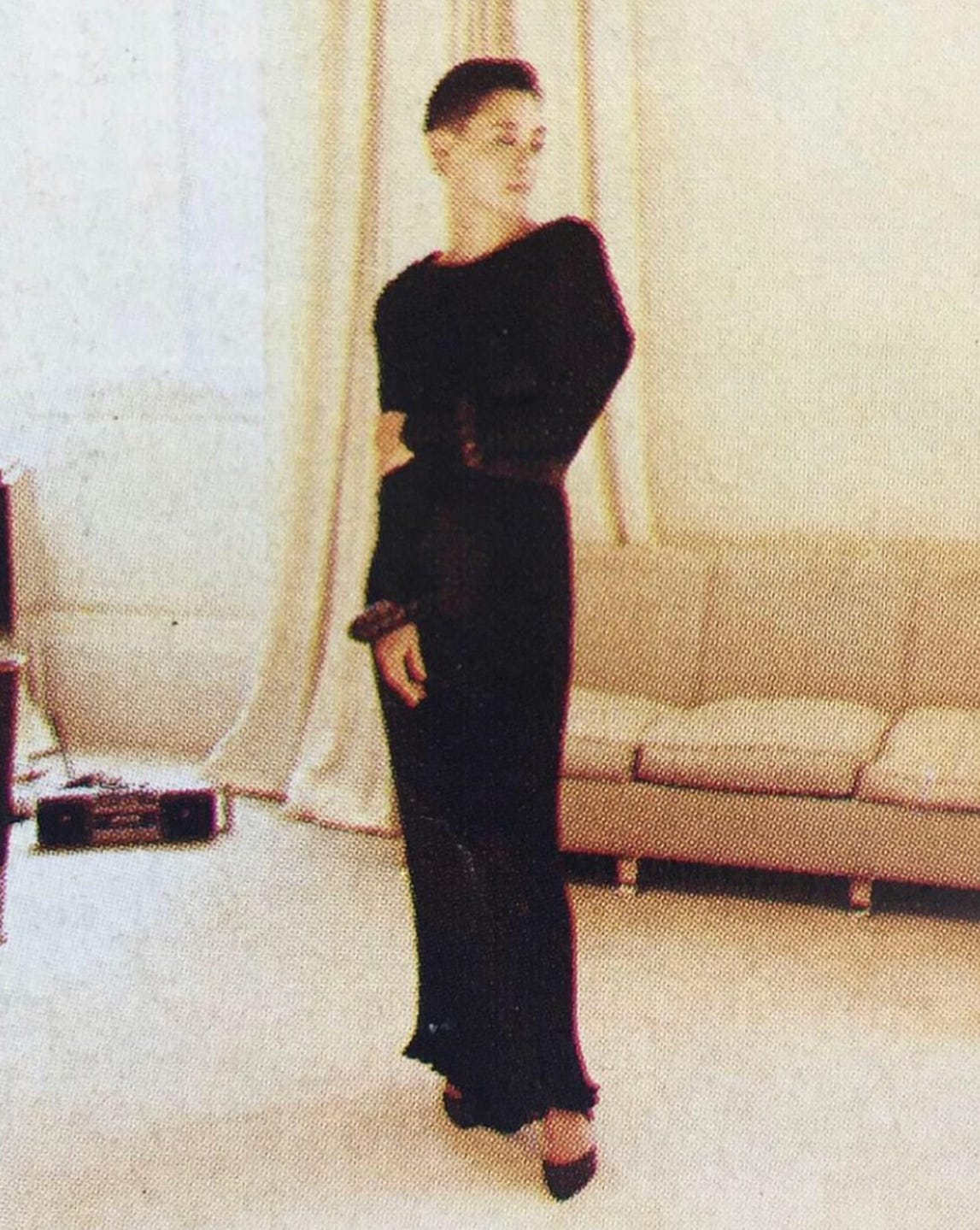 A lone woman with short hair and a sleek black silk dress stands in the middle of a finely decorated living room with all furnishings and window dressings in shades of cream.