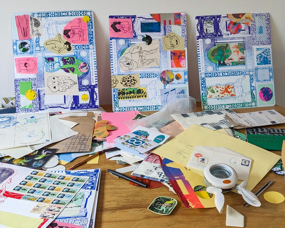 A wooden desk is covered with an assortment of papers, vintage stamps, and art supplies, creating a dyanmic workspace. Three mixed-media collages featuring blueprints, hand-drawn figures, and colorful cutouts lean against the wall. 
