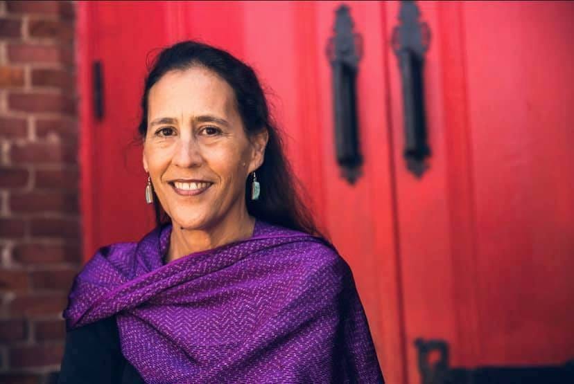 Julie looks directly at the camera, smiling. She has long dark hair, pulled back from her face, dangling earrings, and a woven purple shawl around her shoulders. Behind her is a red double door.