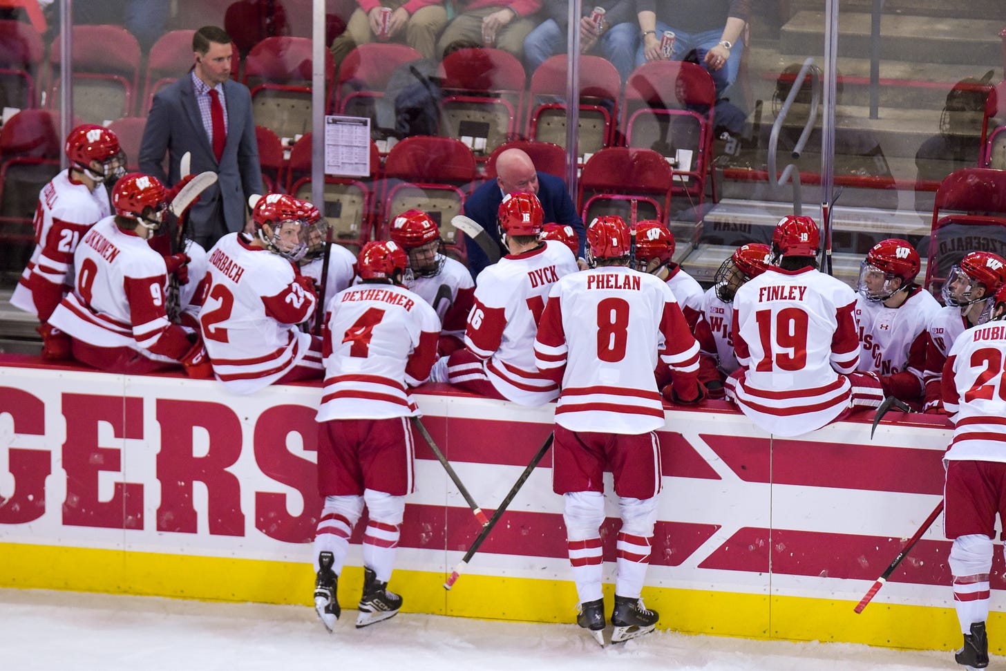 the Wisconsin hockey team sits on the bench, some on the boards, and others stand on the ice while head coach Mike Hastings stands behind the bench speaking to his team