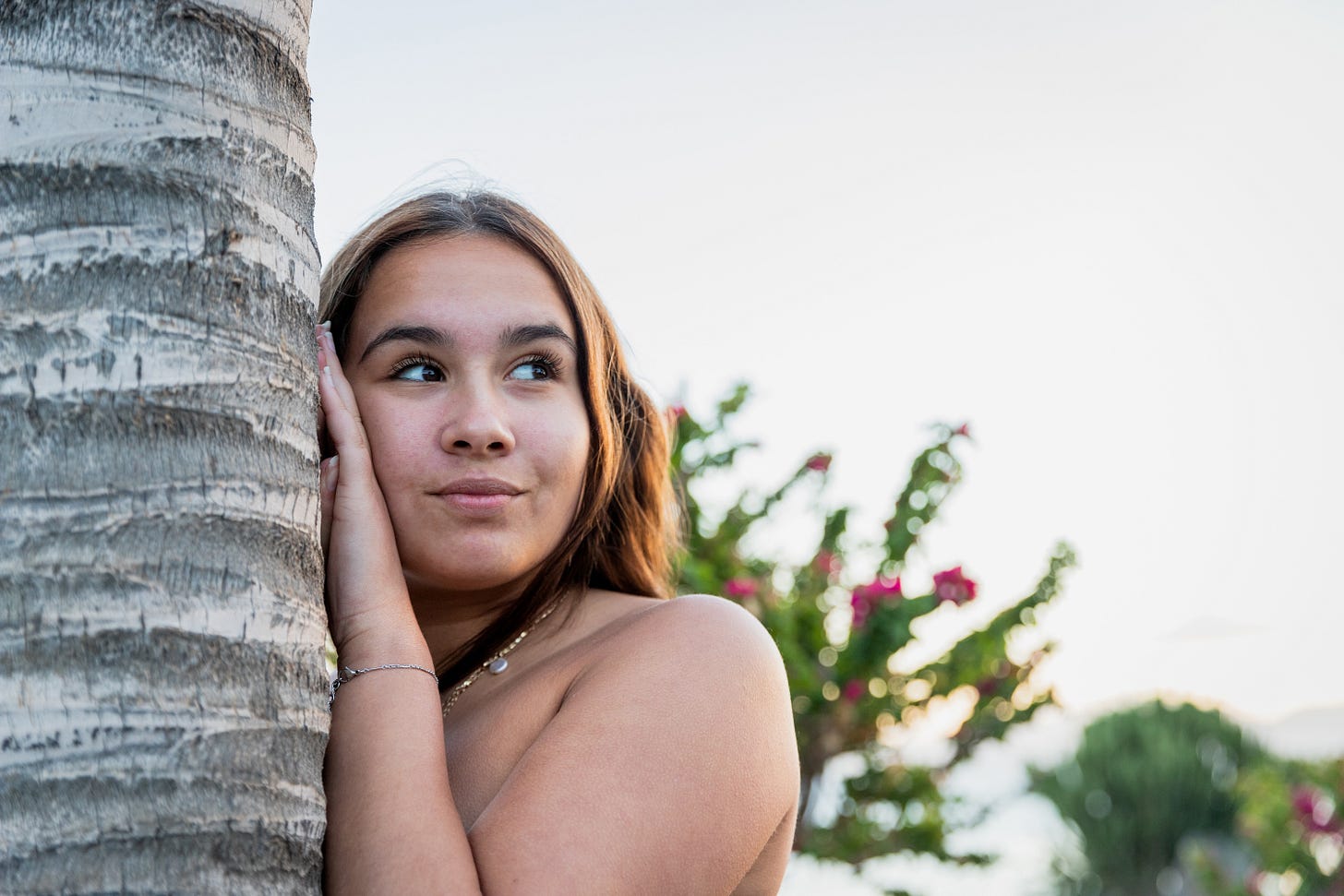 A young woman stands nude behind a palm tree, looking off to the side with a curious, thoughtful expression