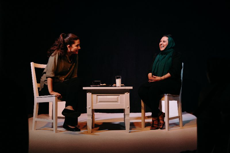 two women sat on white chairs with a white table in the middle. a class of white liquid can be seen on table
