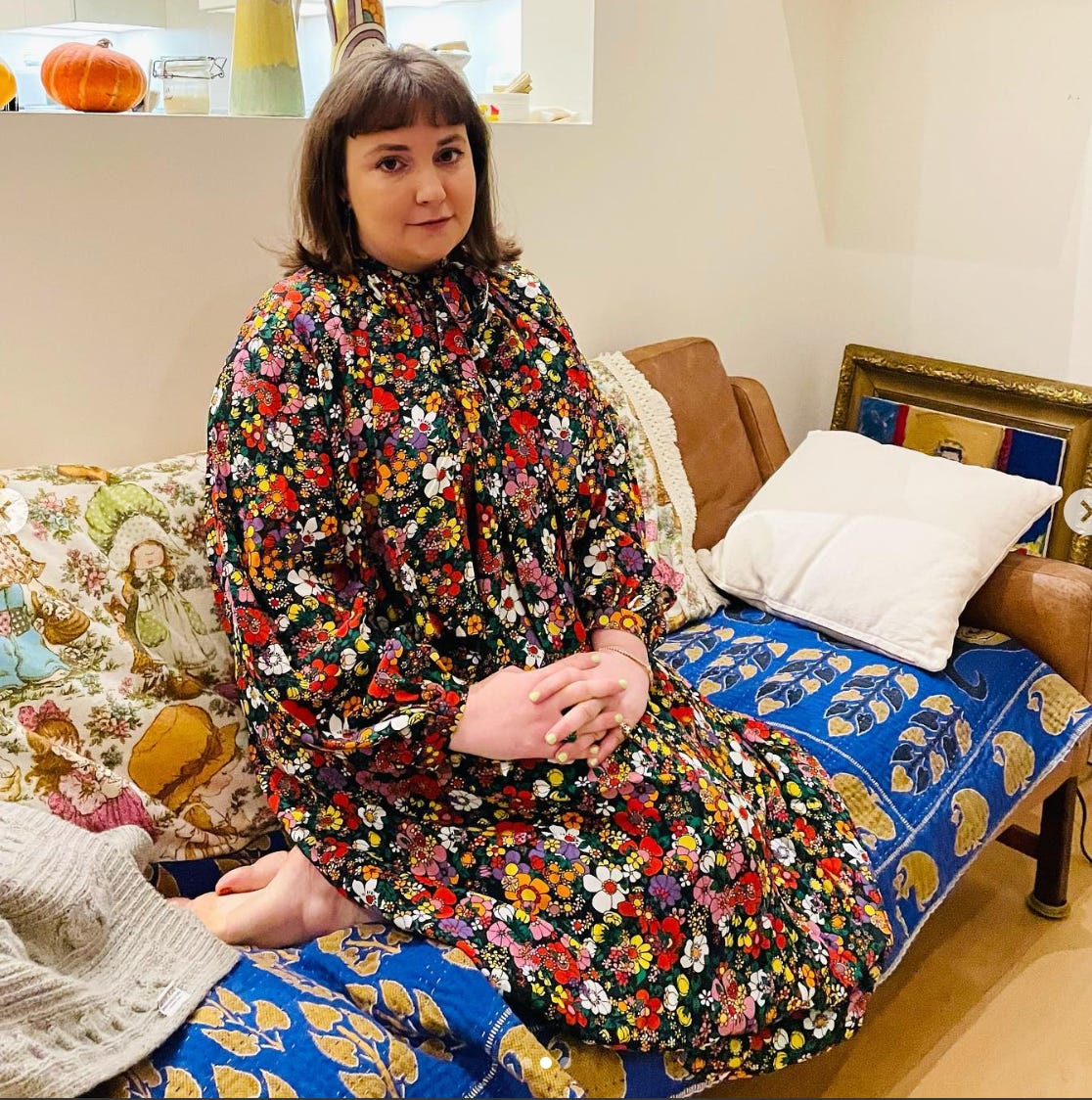 Lena Dunham with a shoulder-length bob in a floor-length flowered dress, sitting on a bed, hands folded over one knee