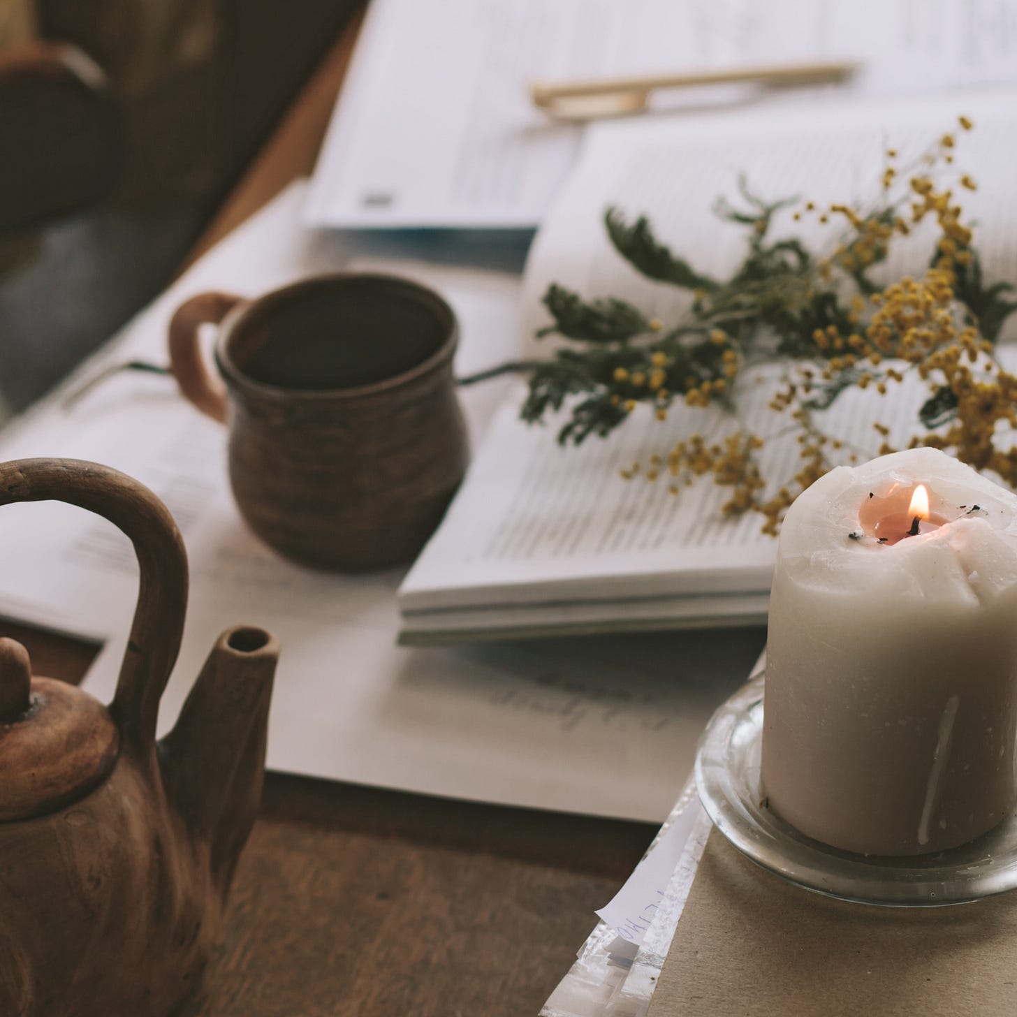 A cosy workspace with a candle, a pottery mug, fresh greenery, and scattered books and papers on a wooden table.