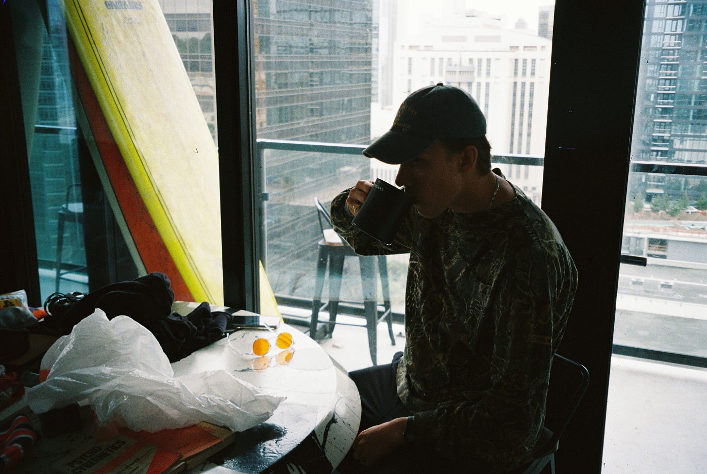 Man dressed in a camo shirt and baseball cap sipping coffee in a backlight scene. Surfboards can be seen on the balcony of the high rise apartment, and a messy table with Psycho-Cybernetics is upturned on the tabletop.