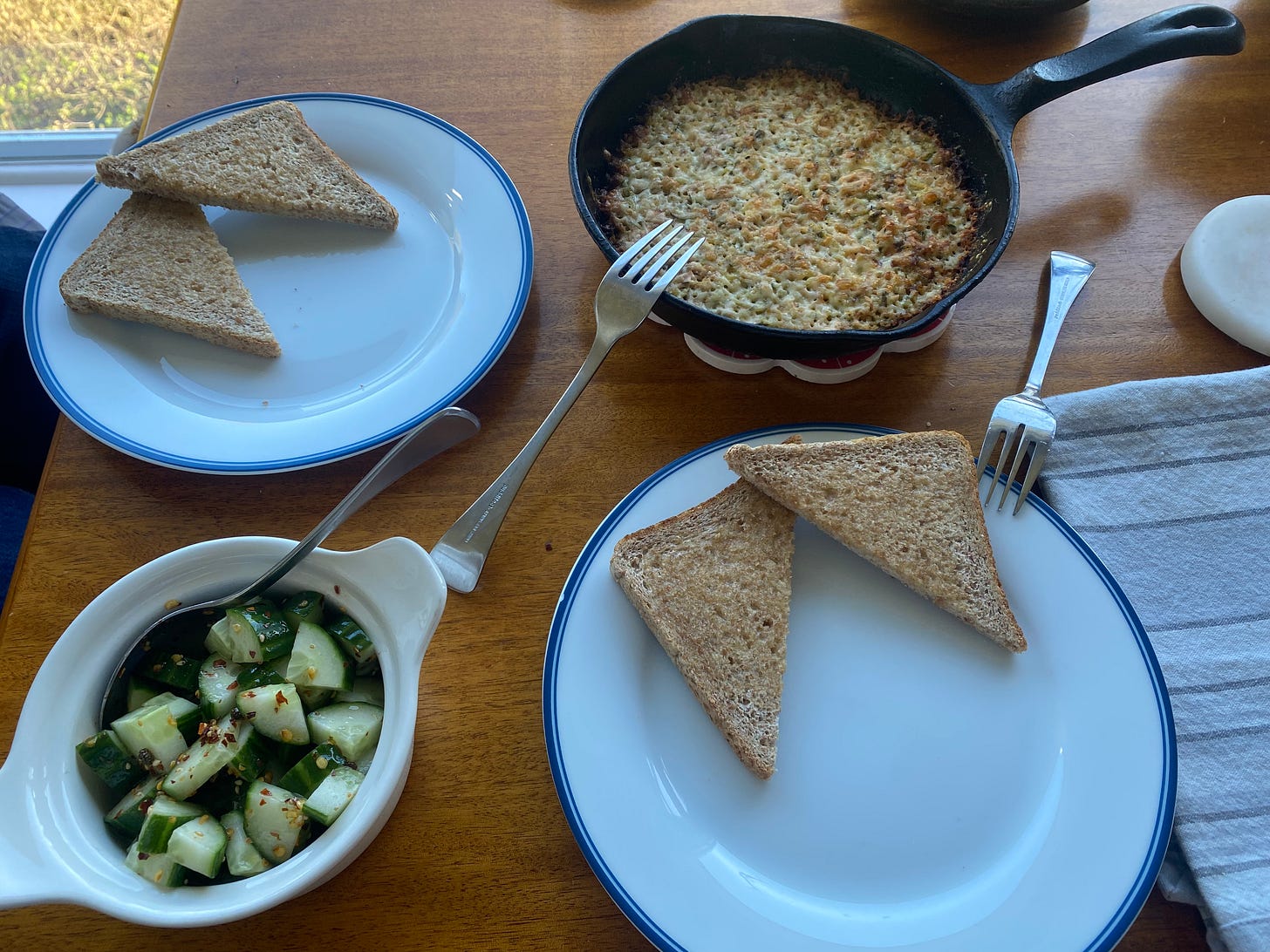 Two plates, each with a halved slice of wheat toast on it. On the table between the plates is a white bowl with the cucumber and chili salad, and a cast iron pan with the tuna melt dish described above. A fork rests on the edge of the pan, and a spoon at the edge of the bowl.