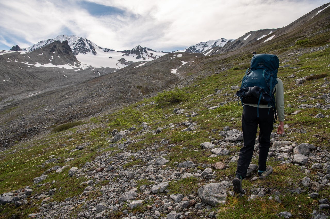 A hiker with a teal pack, seen from behind, walks down a slope of green tundra and scattered pale boulders toward a broad valley. Ahead, rubble-covered slopes rise to snow-streaked peaks and a glacier under a bright, mostly cloudy sky.