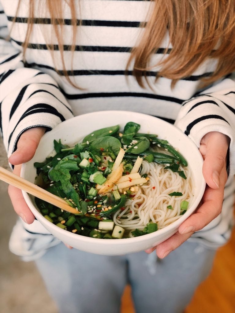 woman in a striped shirt holding a healthy noodle bowl with greens