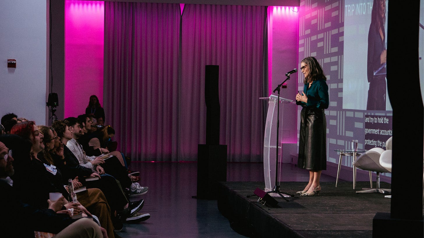 A speaker stands on stage at a podium addressing an audience seated in rows. The room is lit with purple and pink lighting, and a large screen behind the speaker projects her image and presentation text. The setting appears to be a formal event or conference.