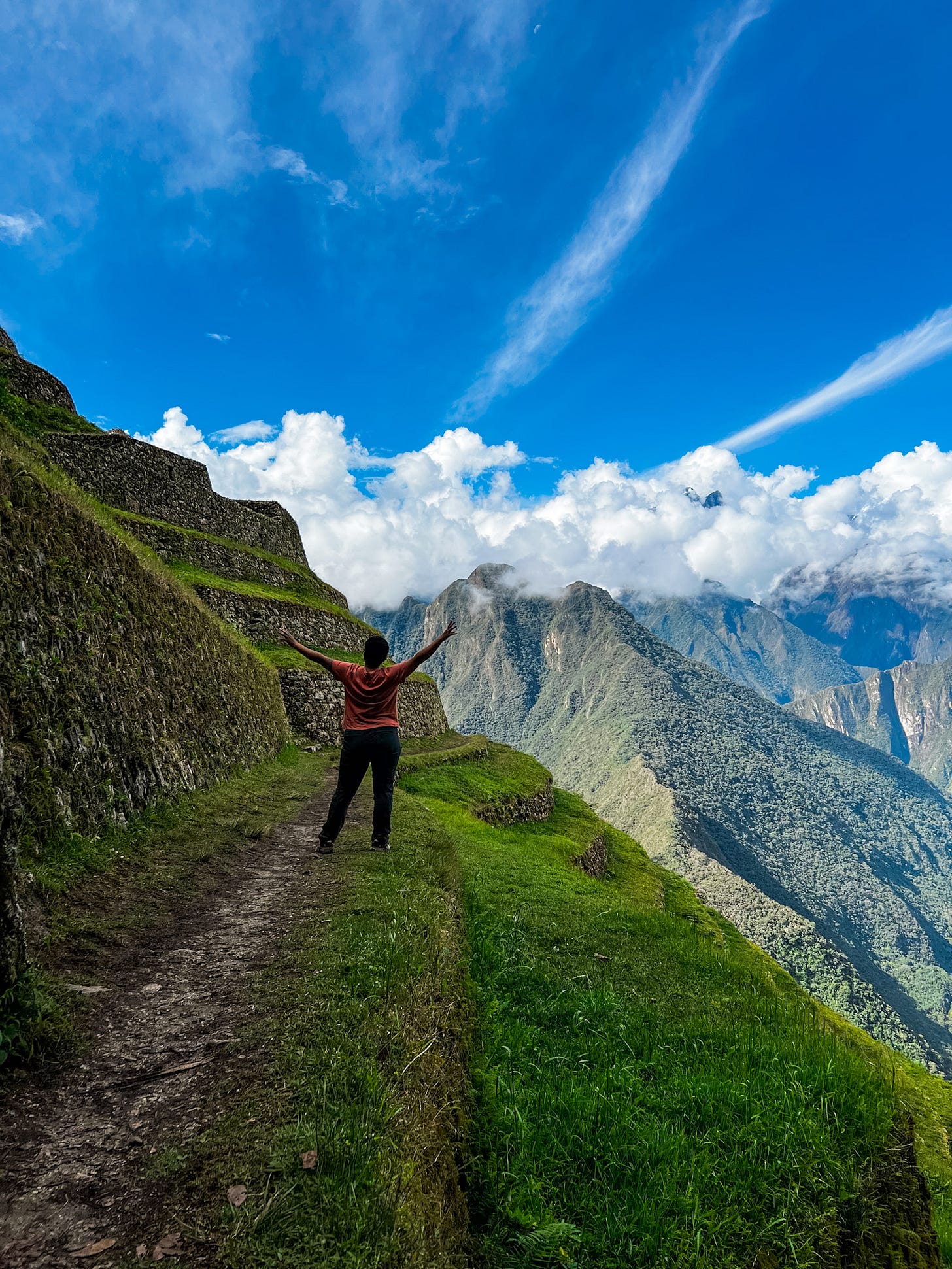 Anela stands on a graduated step, one of many terraced into the hillside part of a former inca site, high up in the mountains with clouds and mountaintops visible behind her with her arms up in the air.