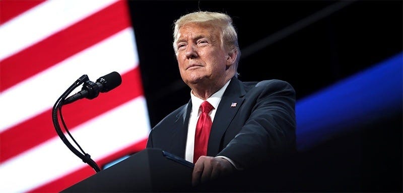 Donald Trump in a dark suit and red tie speaks at podium with microphone. Backdrop shows partial American flag stripes on left, blue lighting on right. Speaker has stern expression, looking off-camera. Image conveys political speech or rally atmosphere. Donald Trump in a dark suit and red tie speaks at podium with microphone. Backdrop shows partial American flag stripes on left, blue lighting on right. Speaker has stern expression, looking off-camera. Image conveys political speech or rally atmosphere.