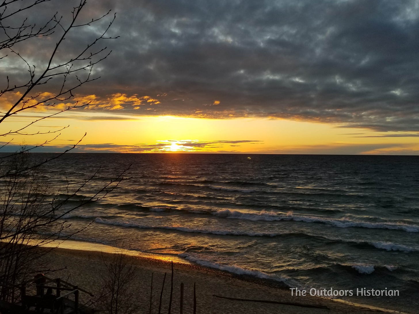 A setting sunset creates a golden sky that reflects on a wavy Lake Superior with white caps crashing along a beach.