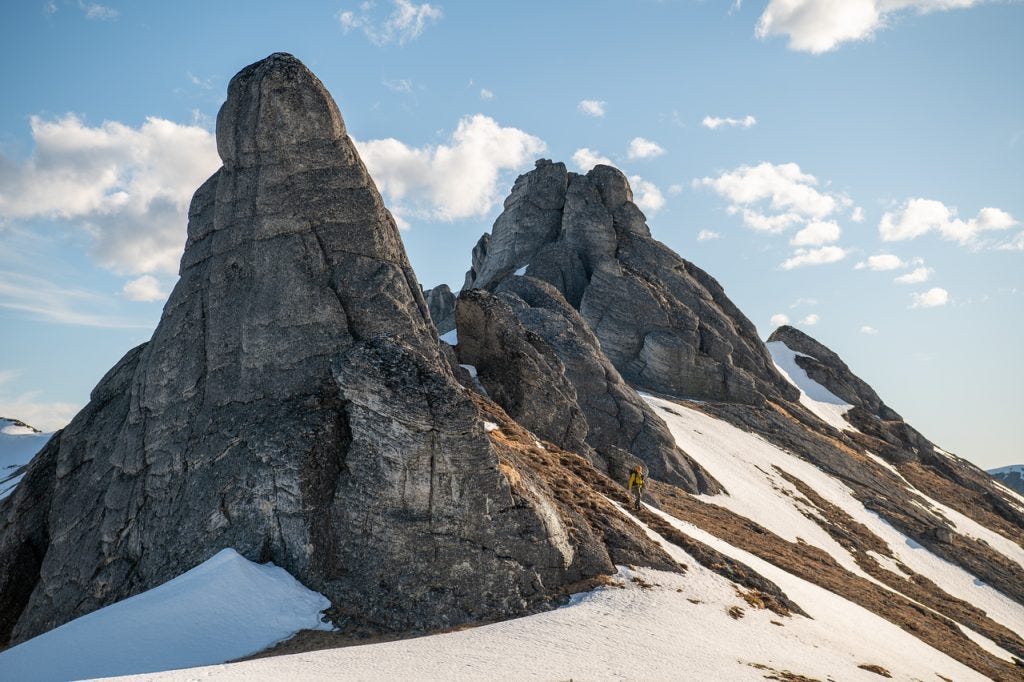 Tall granite spires tower over a tiny figure in a yellow jacket standing at their base, a snow slope below and blue sky above, the small figure making the rock's scale clear.