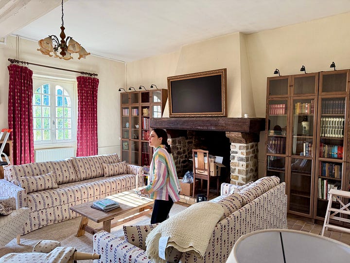 A woman walking through a furnished coach house sitting room; an open cupboard of folded white linens and towels; a woman in a straw hat clipping hydrangeas; a blue-and-white vase of pink and purple hydrangeas.