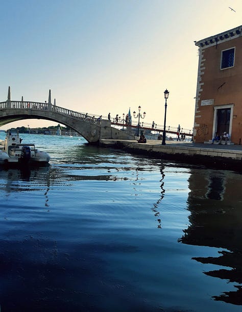 Image 1: A picturesque stone bridge crossing a Venetian canal at sunset, with people walking across and reflections of historic Italian architecture shimmering in the blue water. A small boat is docked on the left, and a terracotta building with white trim stands on the right. Photo taken in Venice, Italy. Image 2: Outdoor summer gathering in an Italian backyard, featuring a plastic cup with a pink cocktail in the foreground, palm trees, and a group of friends playing beer pong in the background. The sun is shining behind the trees, giving a relaxed Italian party vibe. Photo taken in Italy. Image 3: Classic Venetian canal scene with colorful historic buildings, a moored boat, and calm water reflecting the architecture. The canal stretches toward a distant clock tower under a bright blue sky. Photo taken in Venice, Italy. Image 4: Black-and-white photograph of Venetian canal houses and rippling water, highlighting the texture and timeless character of Italy’s iconic canal-side architecture. Taken in Venice, Italy. Image 5: Close-up of a gray cat perched on a stone ledge with lush Italian greenery behind, while another cat lounges on the pavement next to a blue car. The evening sky is partly cloudy, capturing a quiet residential moment in Italy.