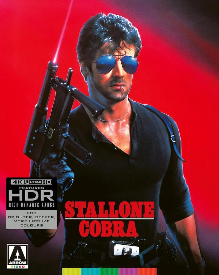 Cobra (1986) Arrow Video 4K UHD cover art with Sylvester Stallone – Stallone as Lt. Marion “Cobra” Cobretti, holding a submachine gun in front of a vivid red background, promoting the Dolby Vision HDR release.