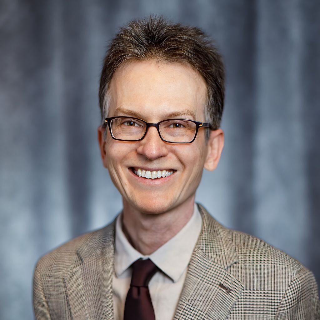 Jay Pea wearing a checked blazer, white shirt, and dark red tie against a blue backdrop. Jay Pea wearing a checked blazer, white shirt, and dark red tie against a blue backdrop.