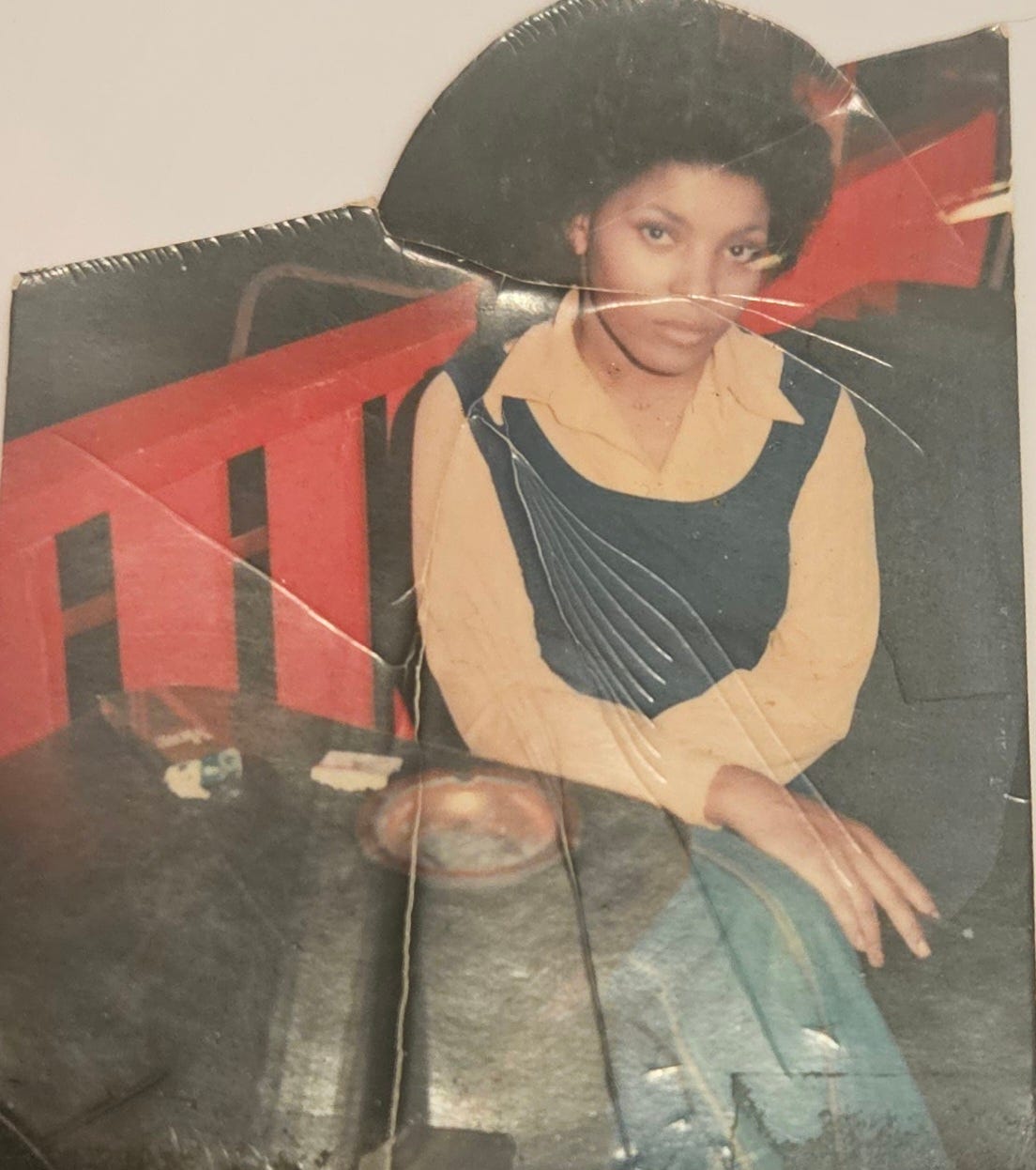 Photo of the author's mother sitting at a table in a restaurant. She's wearing a peach shirt with a butterfly collar under a blue vest. She sits in blue jeans with her legs crossed and holding a More cigarette between her fingers. Her expression is simultaneously stoic and inviting. Photo of the author's mother sitting at a table in a restaurant. She's wearing a peach shirt with a butterfly collar under a blue vest. She sits in blue jeans with her legs crossed and holding a More cigarette between her fingers. Her expression is simultaneously stoic and inviting.