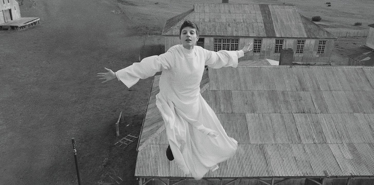 uma mulher branca e de cabelo curto pixie voa pelos ares com umvestido de mangas compridas branco. Ela parece em estado sublime, olhos fechados e braços abertos, e abaixo dela vemos alguns telhados e o campo