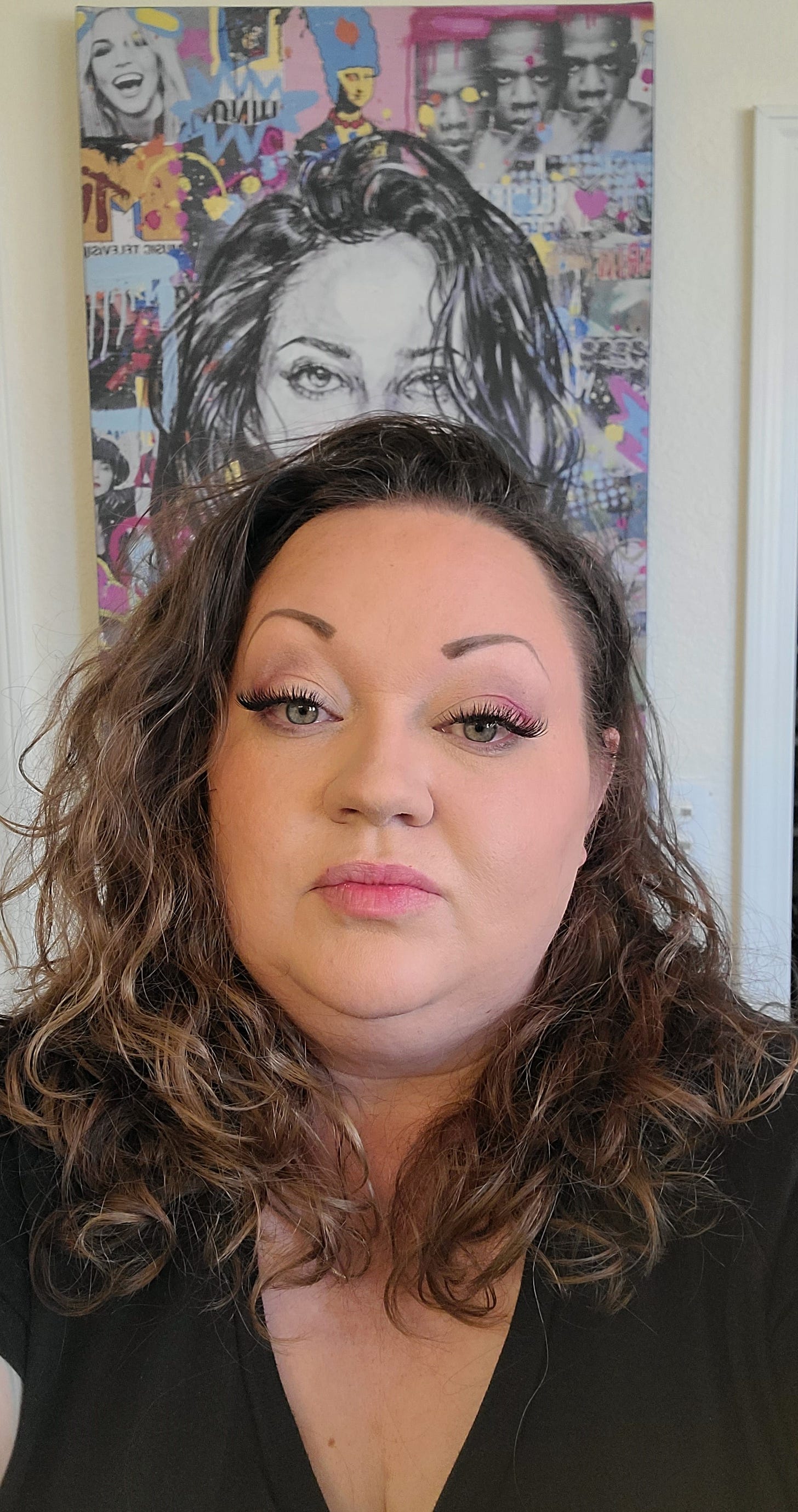 White woman looking directly at the camera, as in a selfie. Her hair resembles (accidentally) a painting behind her of wavy/curly hair, tousled and atypically voluminous, hitting the shoulders and tucked behind one ear. She is wearing a black v-neck tee and has a weird sultry look on her face, but it's working.