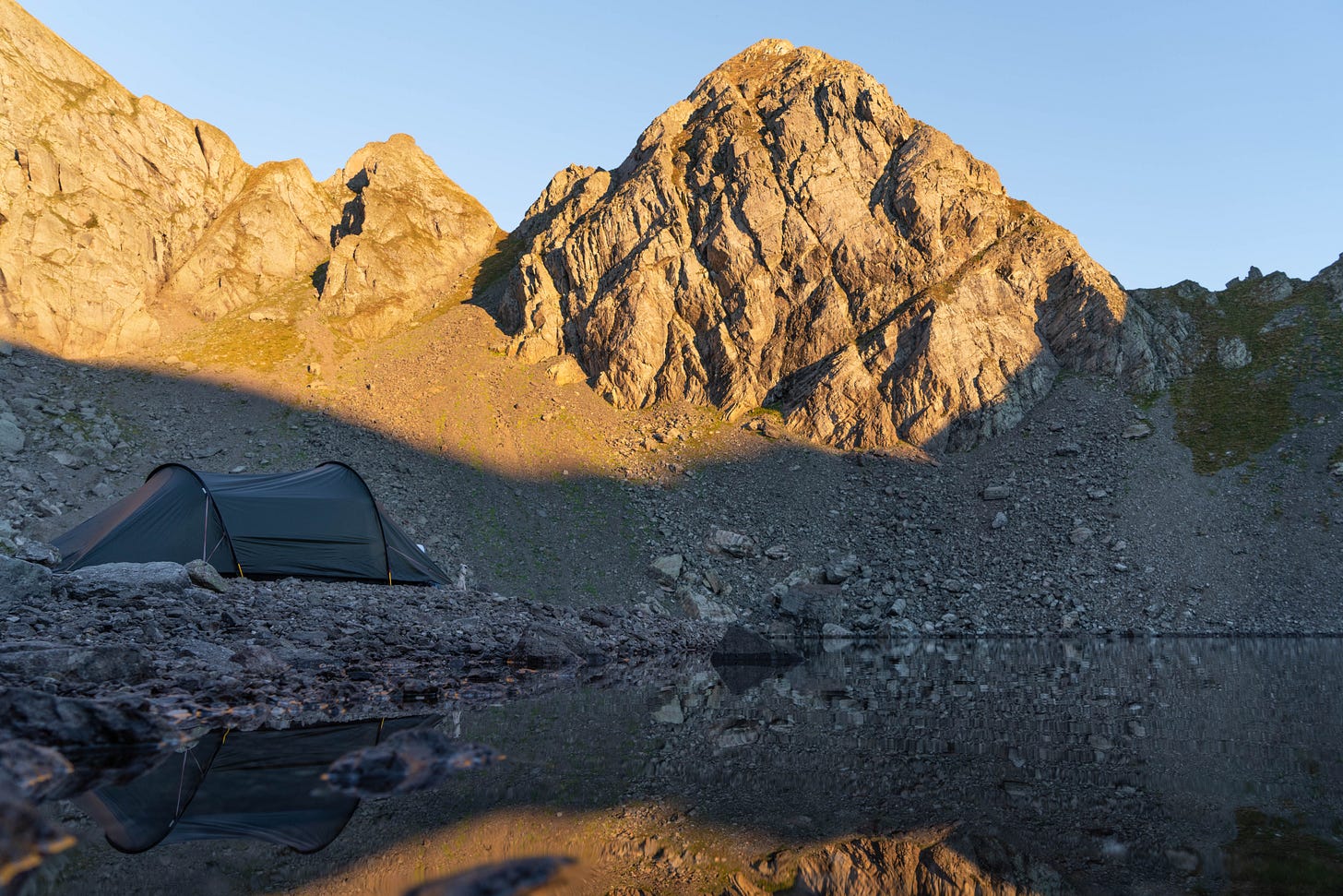 A small green tent blends into the rocky alpine landscape beside Lago Rotondo, a glacial lake at 2,500 metres. The evening light casts long shadows, and the terrain is bare and dramatic. The tent offers a lone pocket of comfort and shelter in the high-altitude wilderness.