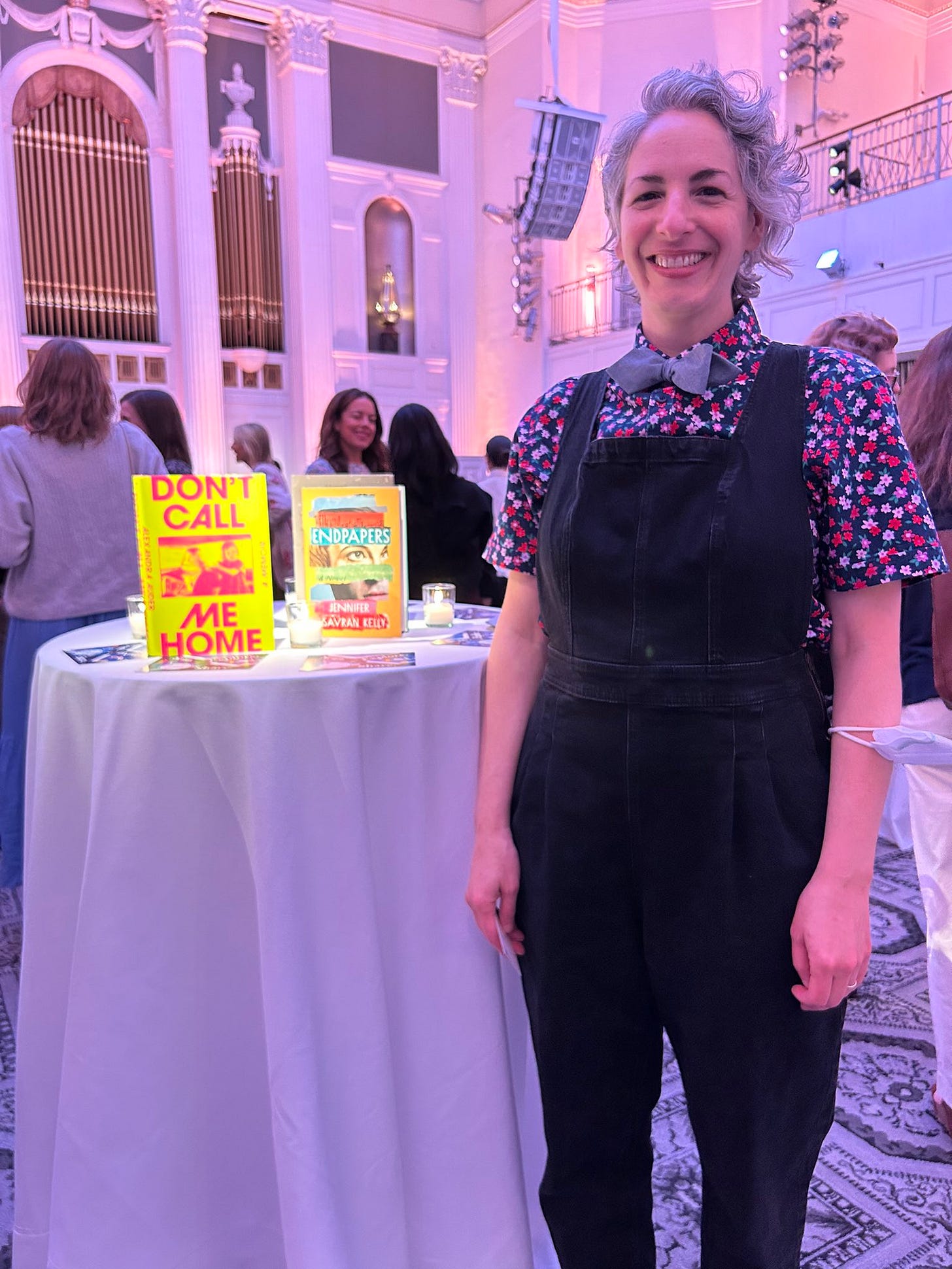 I'm standing next to a small round table that has two books standing up on it. One of them is my novel Endpapers. I'm wearing a jumpsuit with a shortsleeve shirt and bowtie, and I'm smiling.
