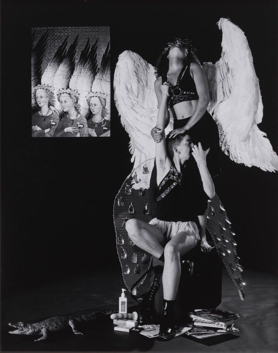 Black and white photo: two figures with angel wings, one standing and holding the other's arm. Books, pamphlets, a faux crocodile, a dildo, and a lotion/lube bottle at their feet. A painting of 3 angels on the left wall adds a surreal tone. The standing angel is the angel with large white wings from the previous images. She is wearing a black top, black pants, and a leather chest harness that the angel underneath is grabbing. The angel that is sitting is kissing the thigh of the other angel behind her, and she is holding her arm and leg. This angel is wearing latex gloves, a black top with a leather chest harness, and short white shorts, with black ankle boots. Their angel wings are large, dark-colored with metal pieces hanging off of them in several different places.